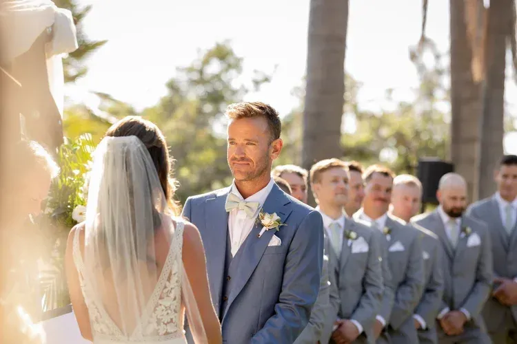 Bride faces groom during outdoor wedding ceremony with groomsmen in gray suits nearby