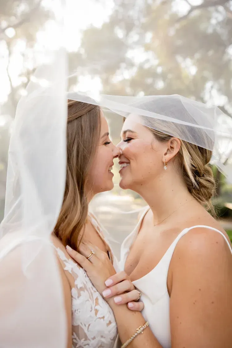 Two brides in white veils share a close, smiling moment outdoors