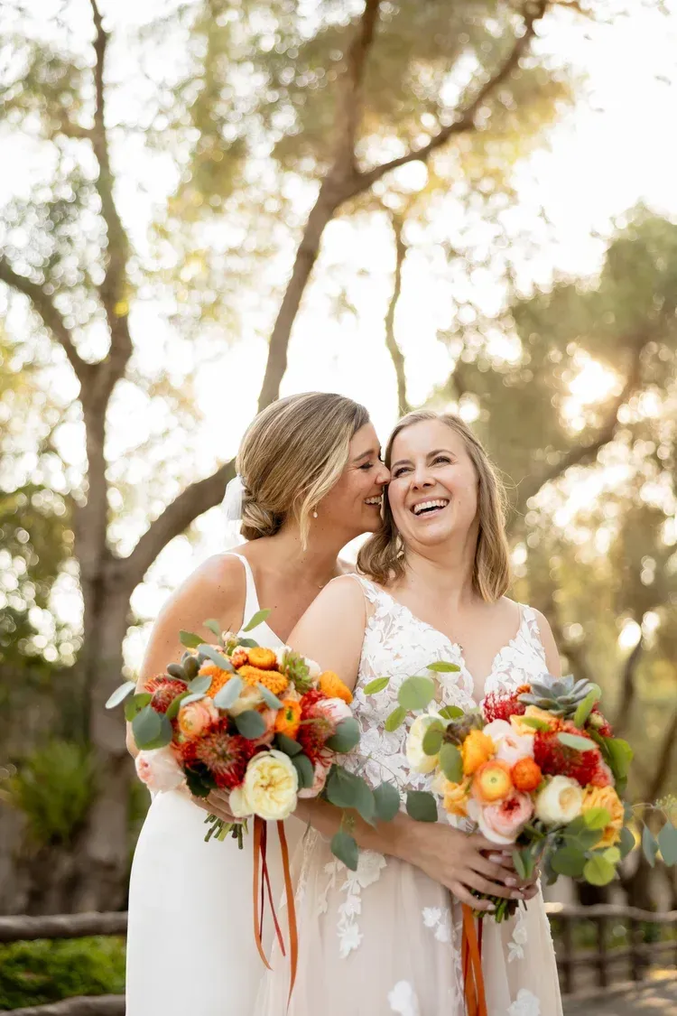 Two brides in white dresses smiling and holding colorful bouquets outdoors at sunset