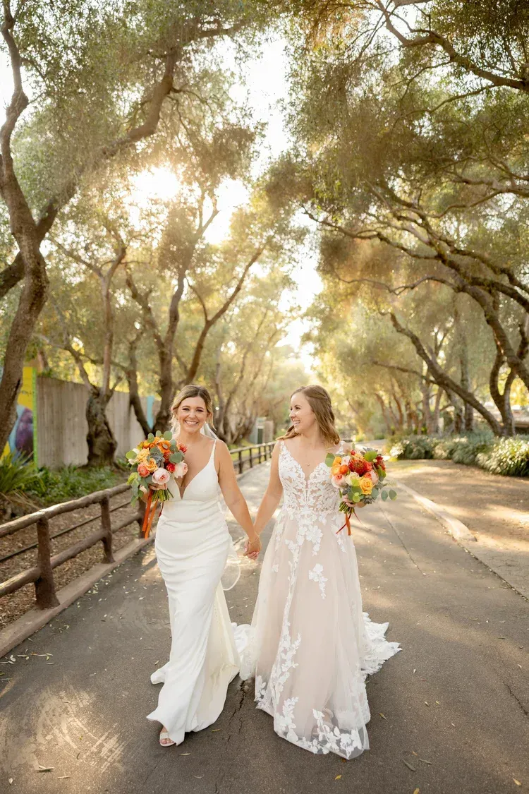 Two brides in white wedding dresses walk hand in hand on a tree-lined road, holding colorful bouquets.