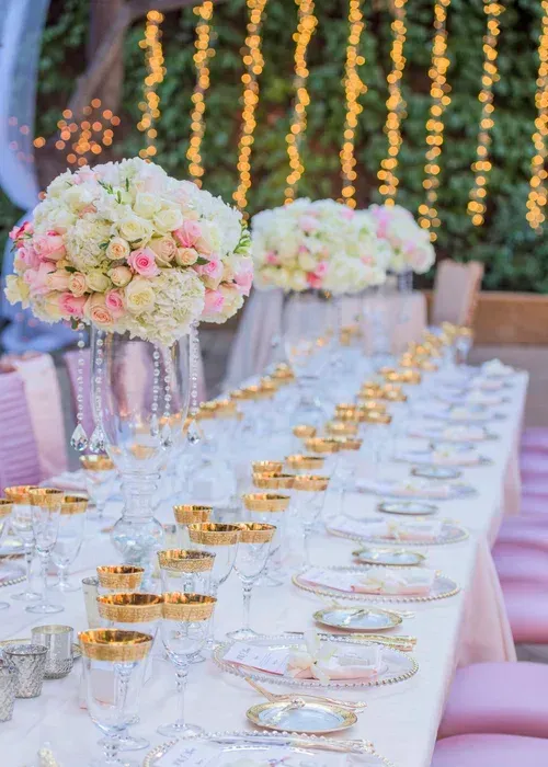 Elegant outdoor reception table with pink floral centerpieces, gold accents, and candlelight at dusk