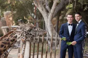 Two men in suits posing beside a giraffe at an outdoor zoo enclosure