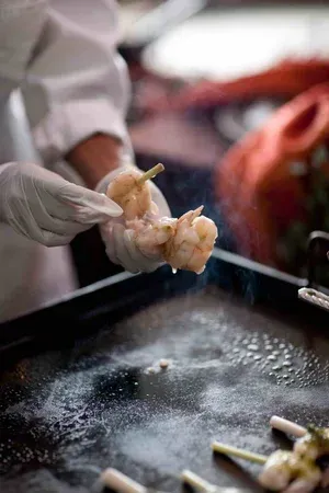 Gloved hands skewer raw shrimp over a steaming hot griddle in a kitchen.