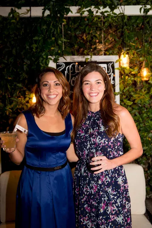 Two women smiling at a nighttime event, one holding a drink, standing before greenery and warm lights.
