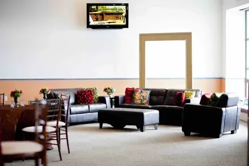 Lobby with black sofas, wooden chairs, potted plants, and a wall-mounted TV.