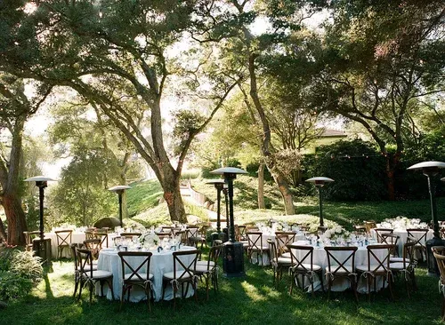Outdoor banquet tables set beneath large trees on a grassy lawn, with white tablecloths and chairs.