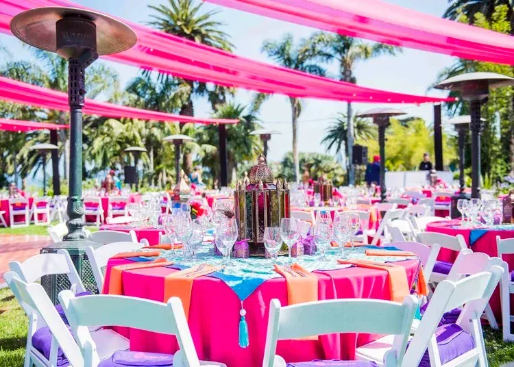 Outdoor event tables with pink tablecloths and white chairs under bright pink drapes, palm trees in the background