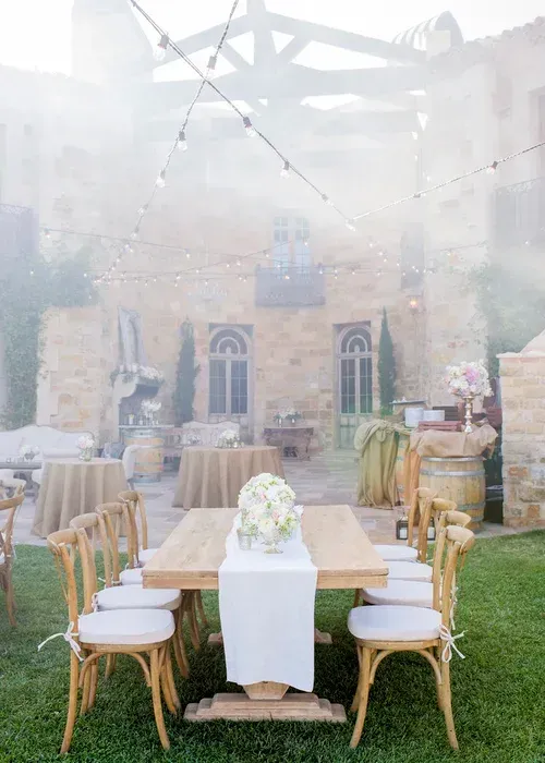 Rustic outdoor dining setup with wicker chairs, long table, and white floral centerpiece in a stone courtyard