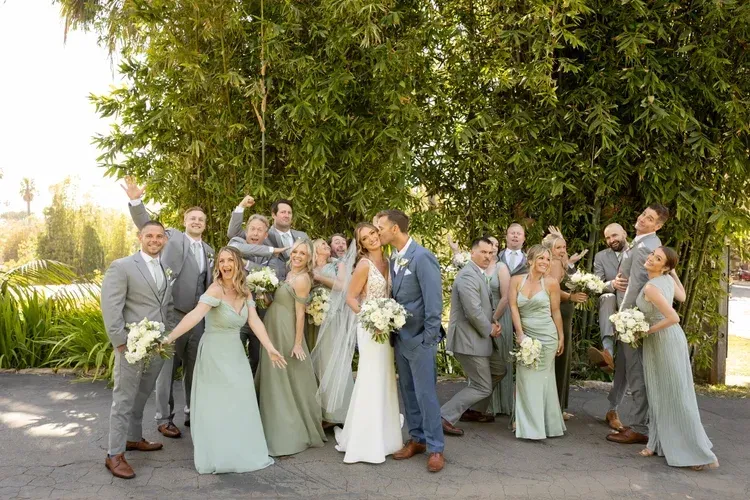 Wedding party posing outdoors in formal attire, with the couple centered in front of greenery.