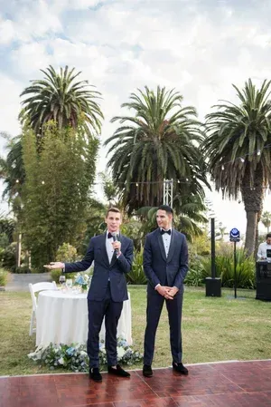 Two men in suits stand outdoors on a lawn with palm trees and a table behind them.