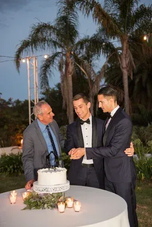 Three men in suits at an outdoor birthday celebration, standing by a white cake on a lit table.