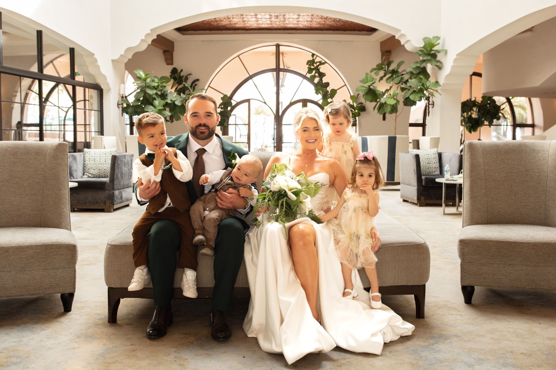 Wedding party seated in a bright venue, with a bride, groom, baby, and young girl holding flowers.