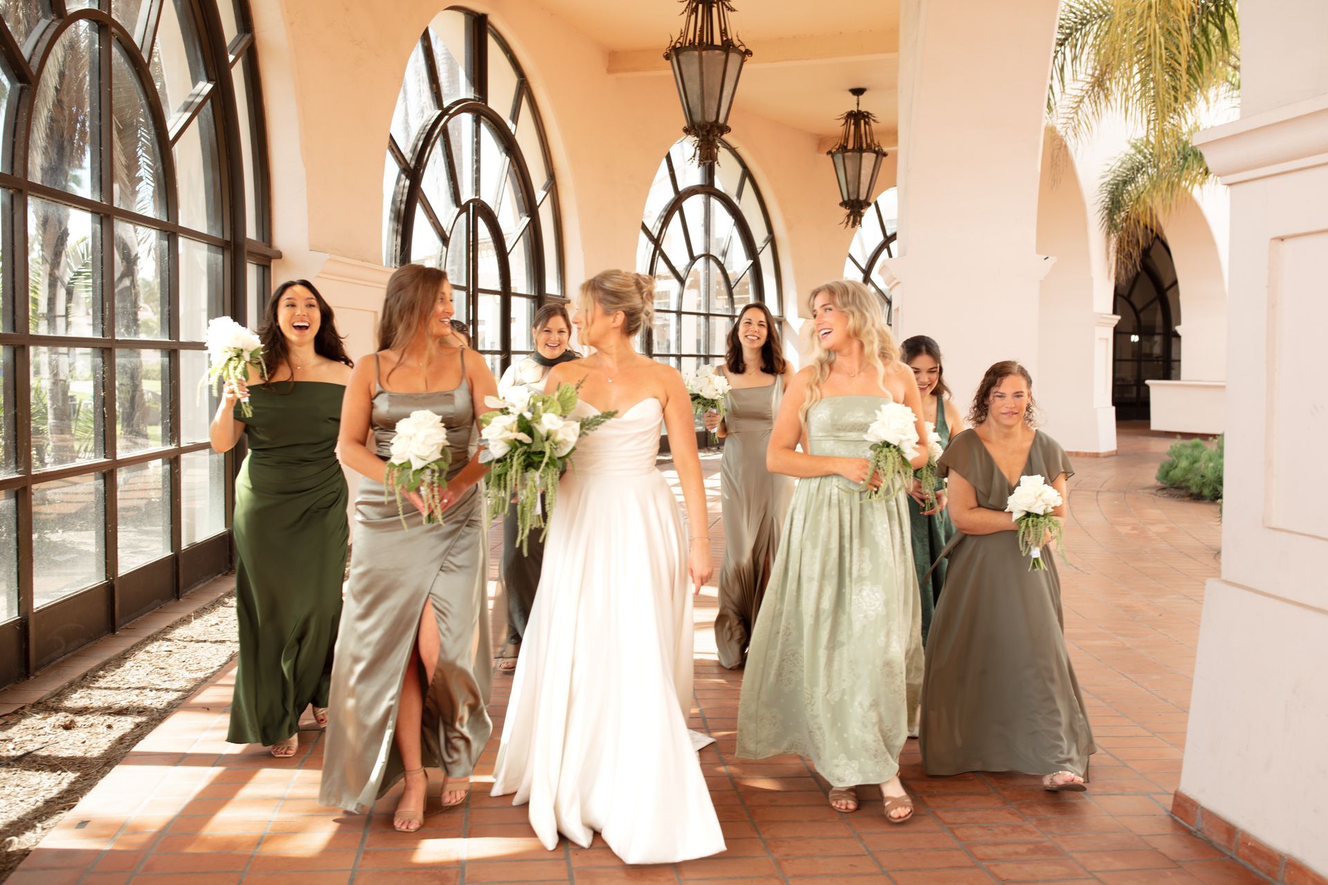 Bridesmaids in green dresses walk with the bride in a white gown through a sunlit archway.