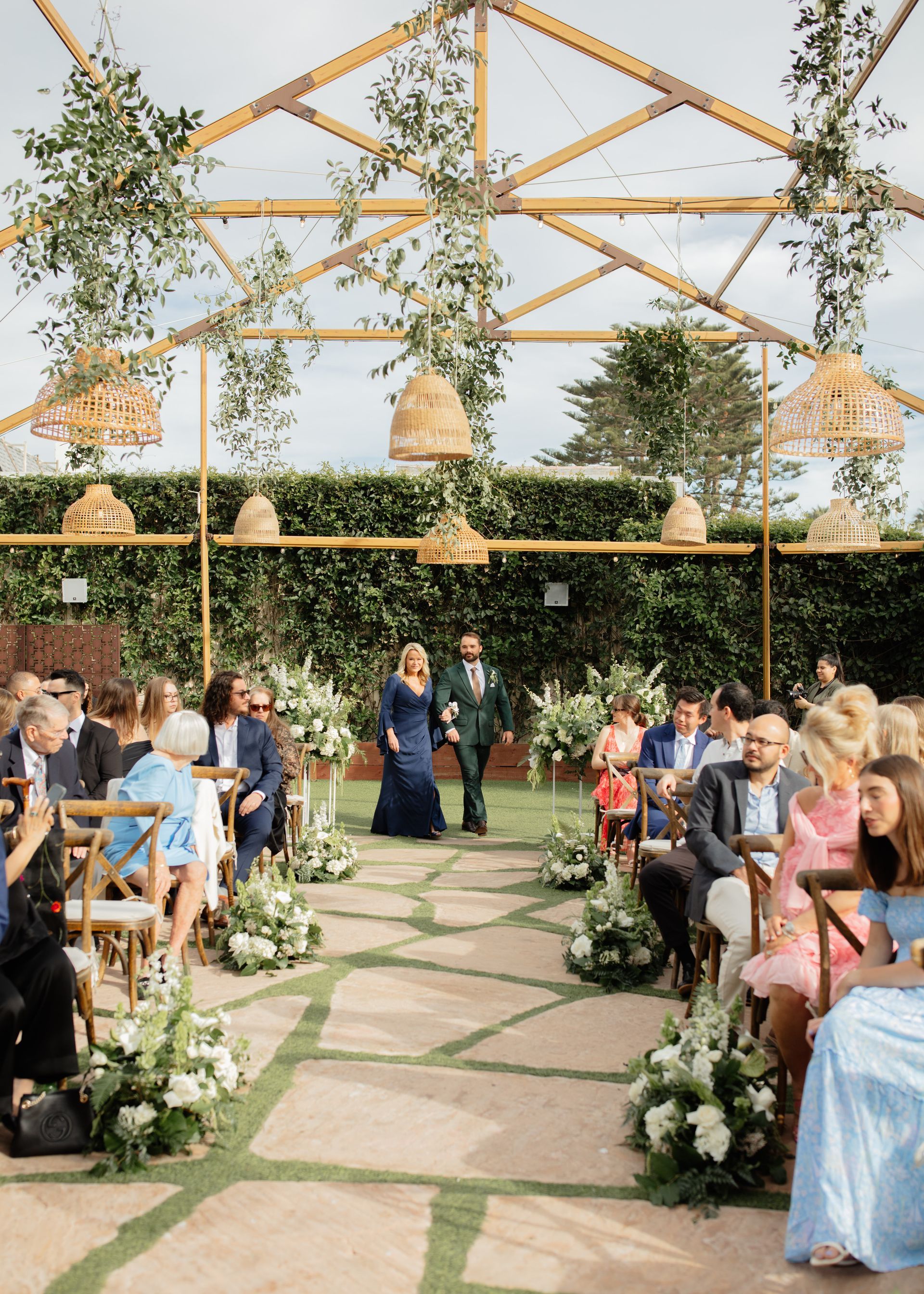 Guests seated along a flower-lined aisle beneath a rustic wooden canopy at an outdoor wedding ceremony
