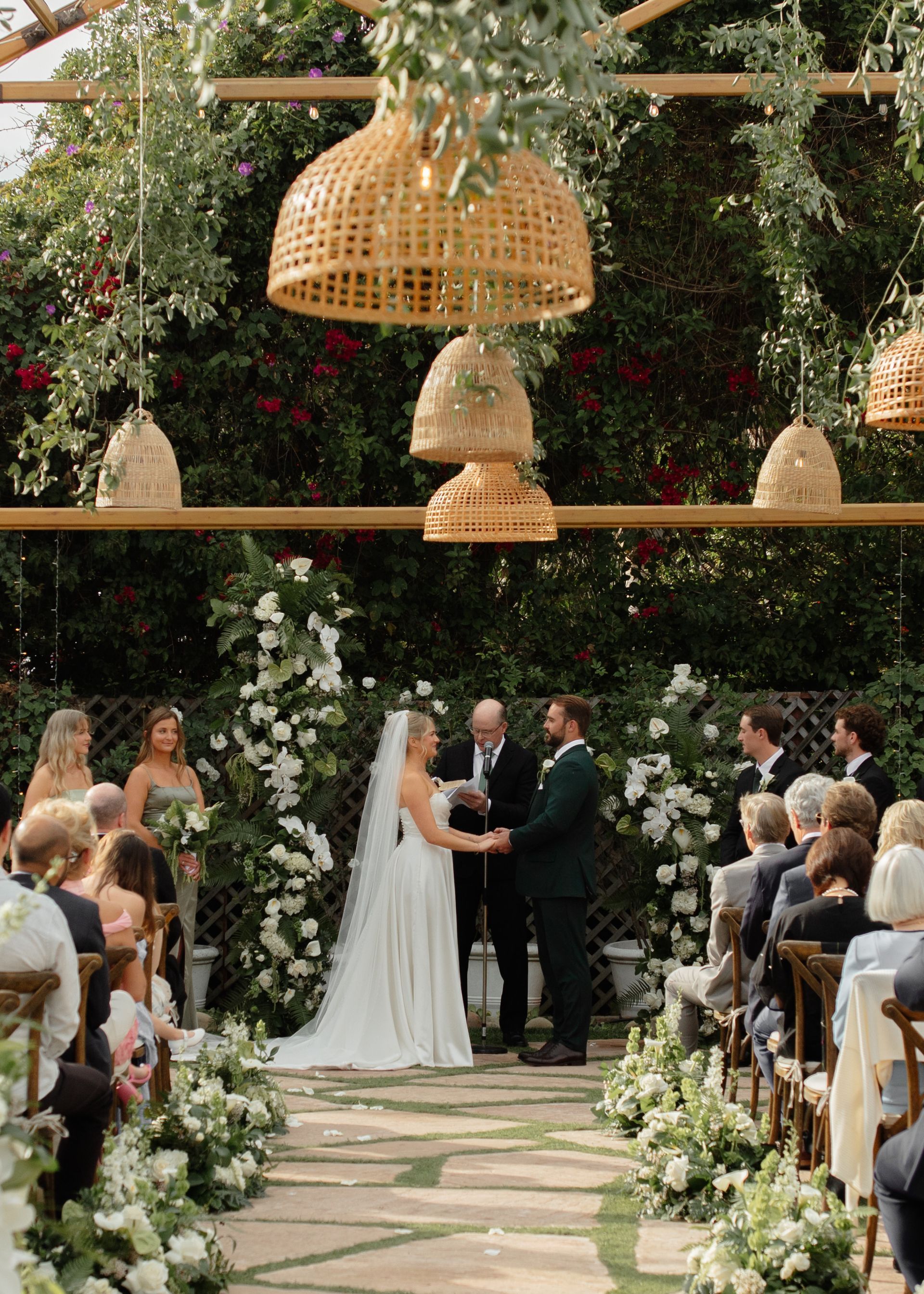 Outdoor wedding ceremony under woven hanging lanterns, with bride and groom exchanging vows in a flower-lined aisle.