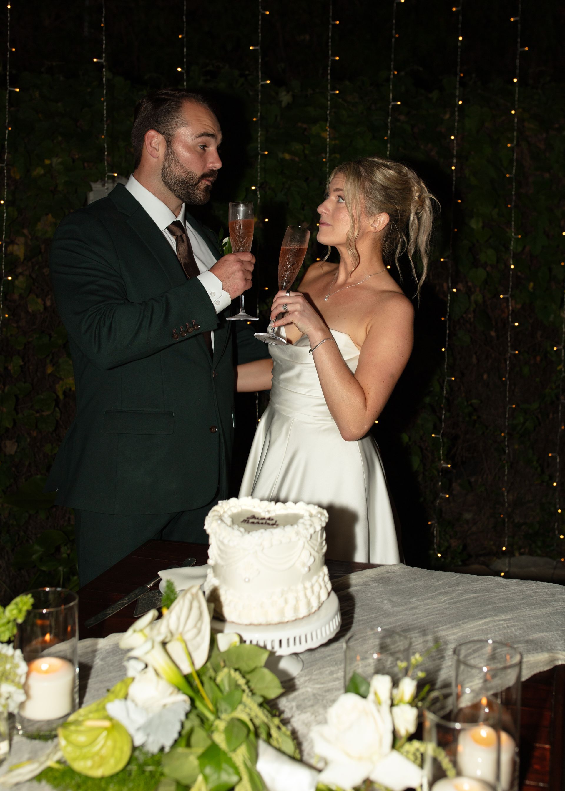 Bride and groom toast beside a white wedding cake at a candlelit reception