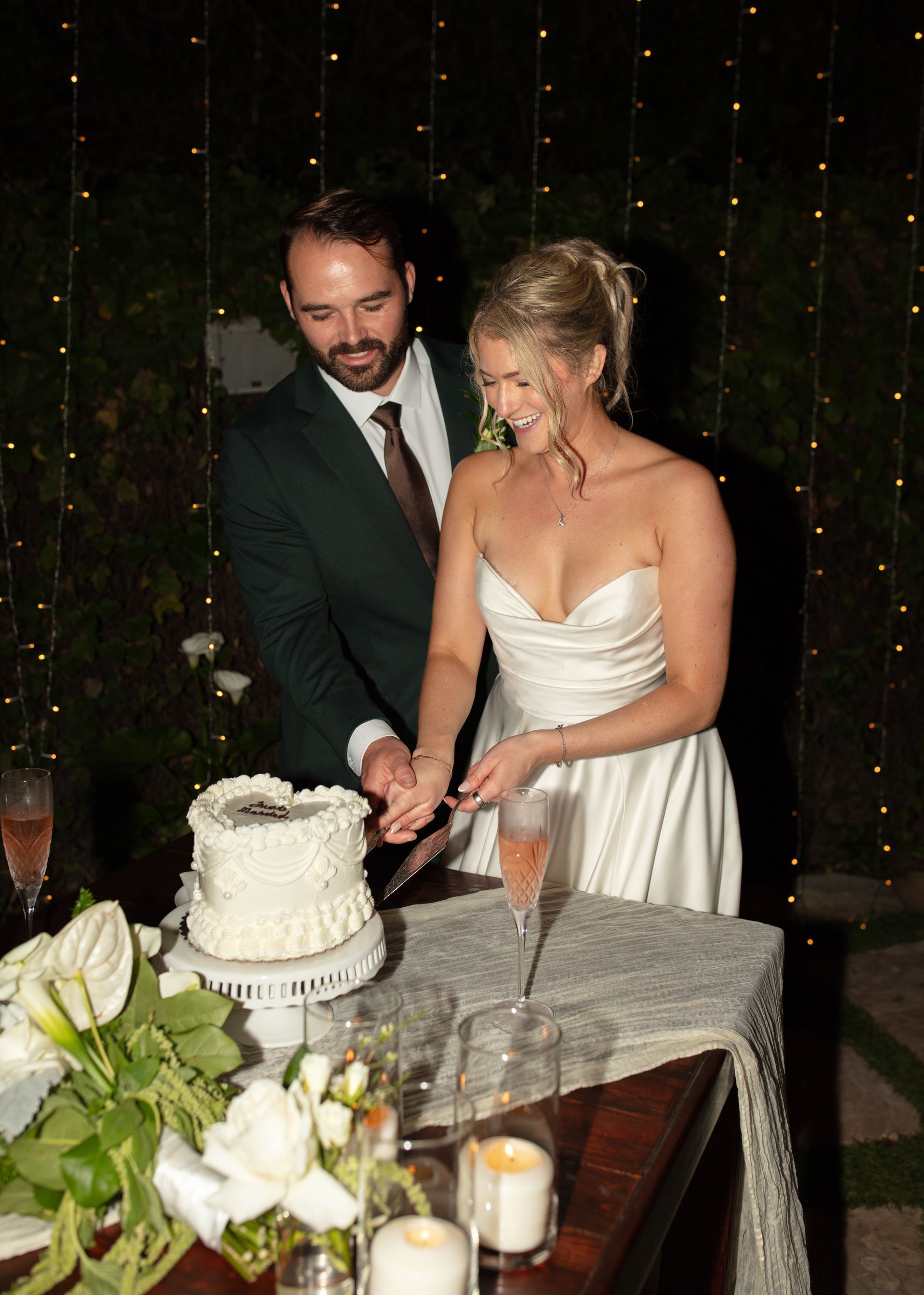 Couple in formal wear cutting a white wedding cake at a reception table.