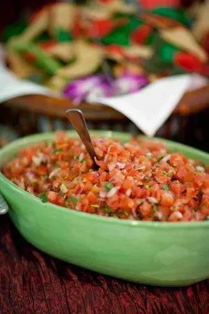 Green bowl of chopped salsa with a spoon, with a blurred tray of snacks in the background