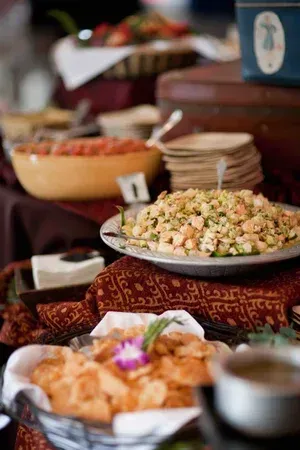 Buffet table with platters of food on patterned red cloth and stacked plates in the background