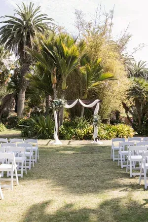 Outdoor wedding ceremony setup with white chairs, an arch draped in fabric, and palm trees in a garden setting