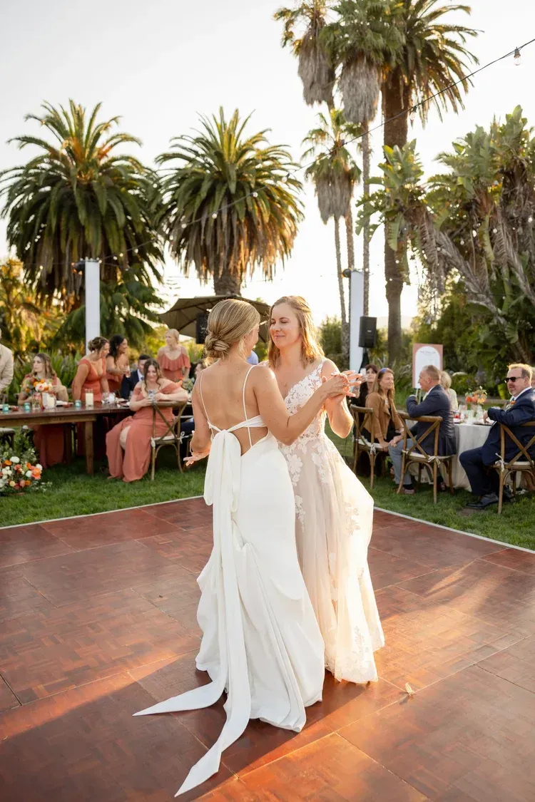 Two people dancing in white dresses on an outdoor terrace at a sunset celebration with palm trees