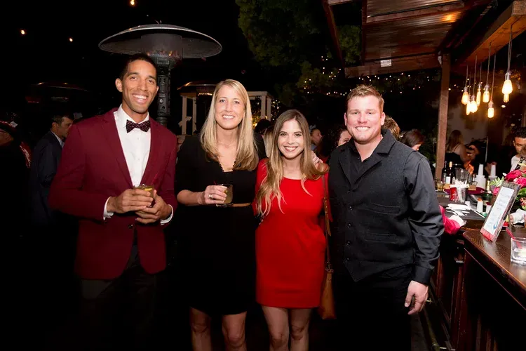 Four people posing with drinks at a dimly lit event bar, wearing formal attire and smiling