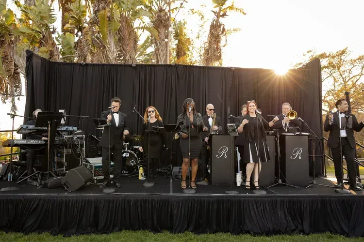 Outdoor band performing on a stage at sunset, with singers, instruments, and sound equipment visible.