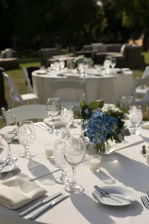 Outdoor banquet tables set with white linens, glassware, and blue floral centerpieces in a garden setting