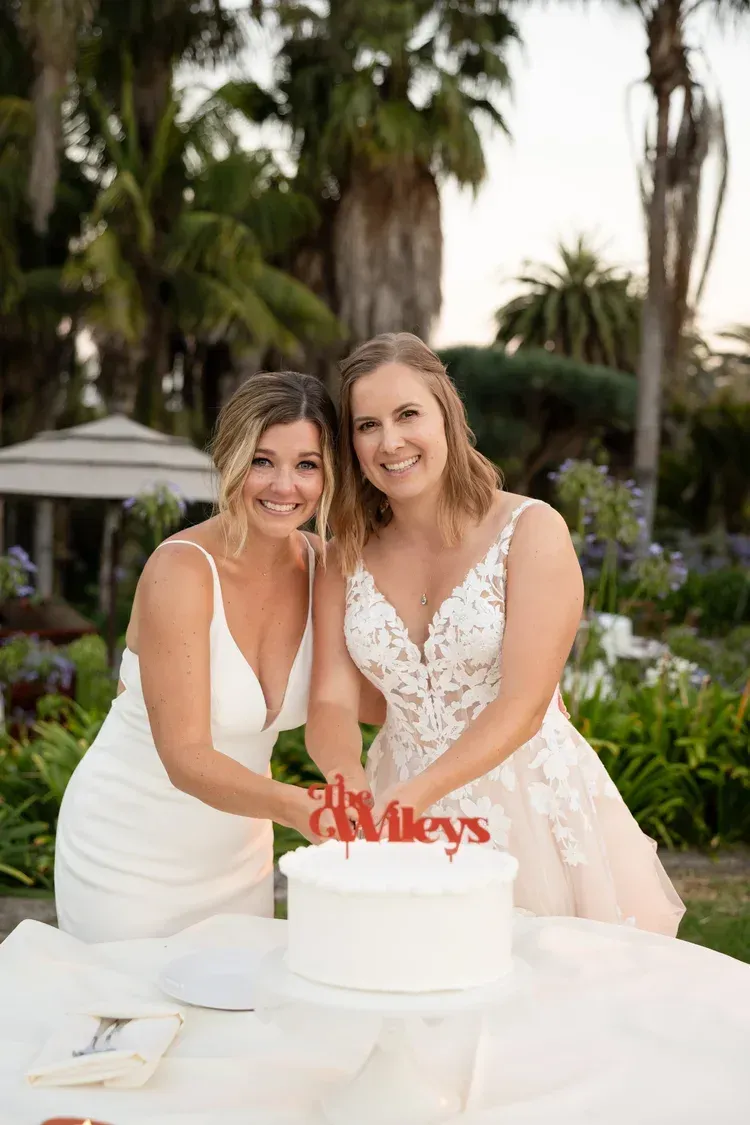 Two women in white dresses smile beside a wedding cake in a tropical garden.