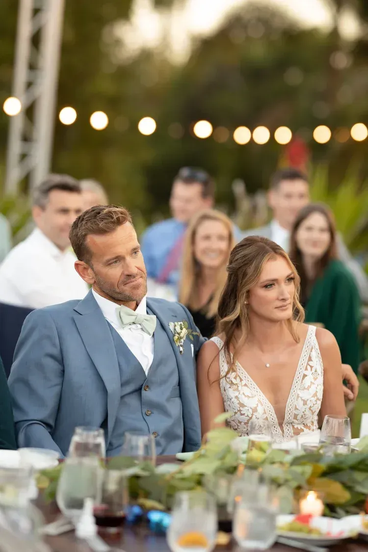 Couple seated at a decorated outdoor wedding reception table with string lights in the background