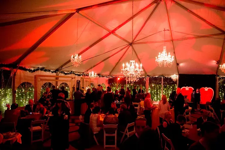 Guests seated under a red-lit tent at a formal event with chandeliers and string lights