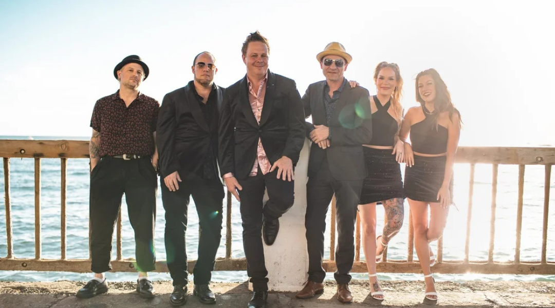 Six people pose on a sunny beach boardwalk with the ocean behind them.