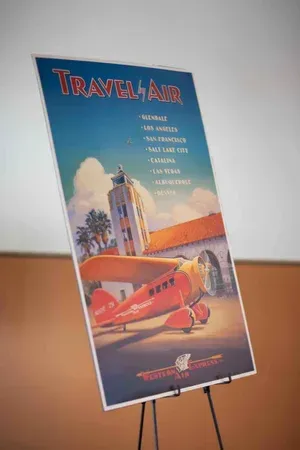 Travel-Air travel poster on a stand, showing a red airplane and a tall building against a blue sky.