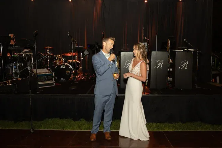 Two people in formal wear talk onstage at a dark event with drums and speakers behind them.