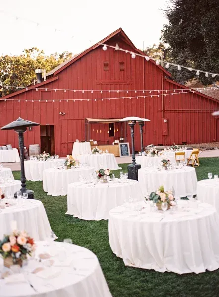 Outdoor wedding reception with white tablecloth tables in front of a red barn