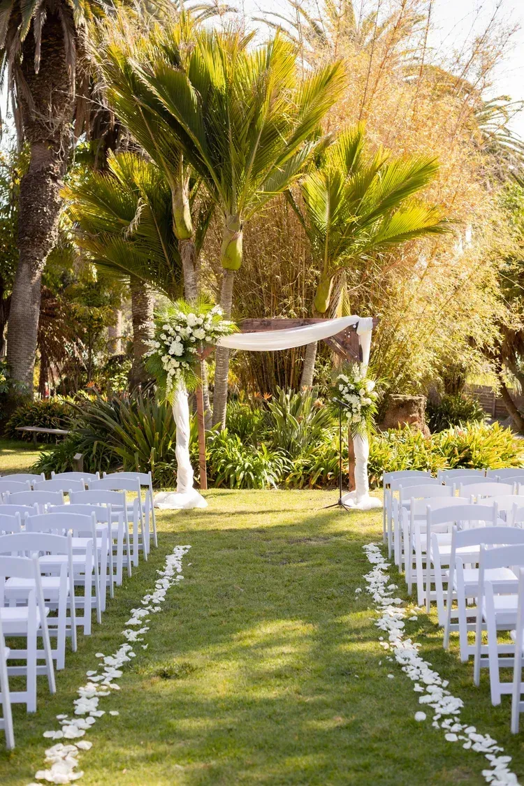 Outdoor wedding aisle with white chairs, flower petals, and a decorated arch under palm trees