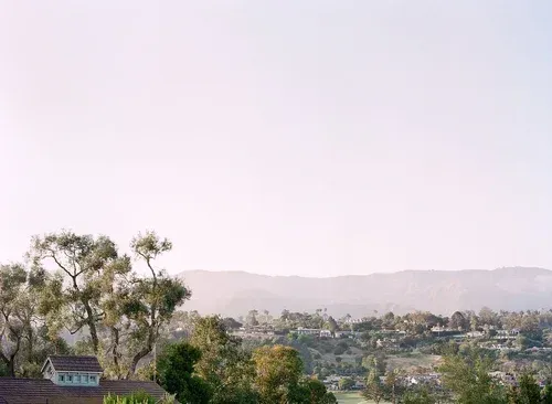 Distant hills above a lush valley at sunrise, with trees and a small house in the foreground.