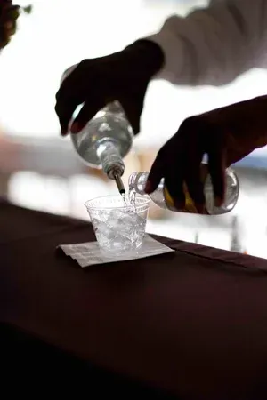 Hands pouring clear liquid into a small cup on a table with a napkin under it