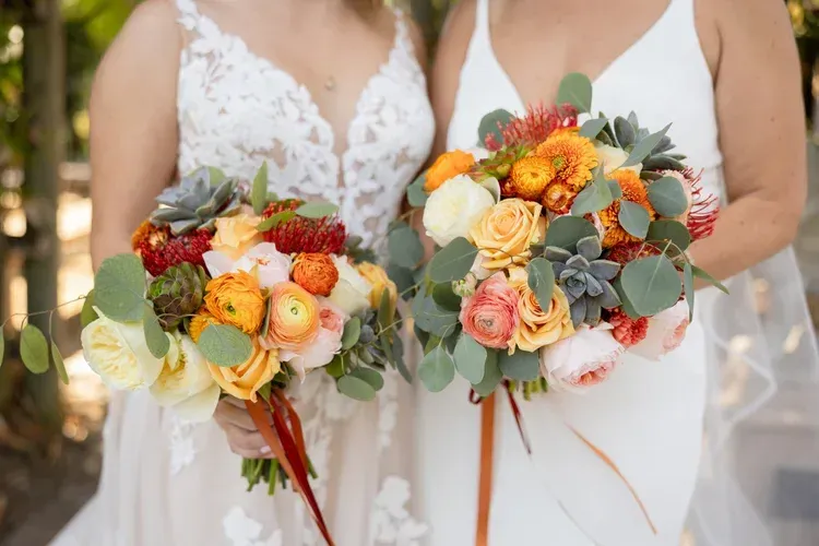 Two brides holding colorful bouquets with roses and greenery, wearing white wedding dresses