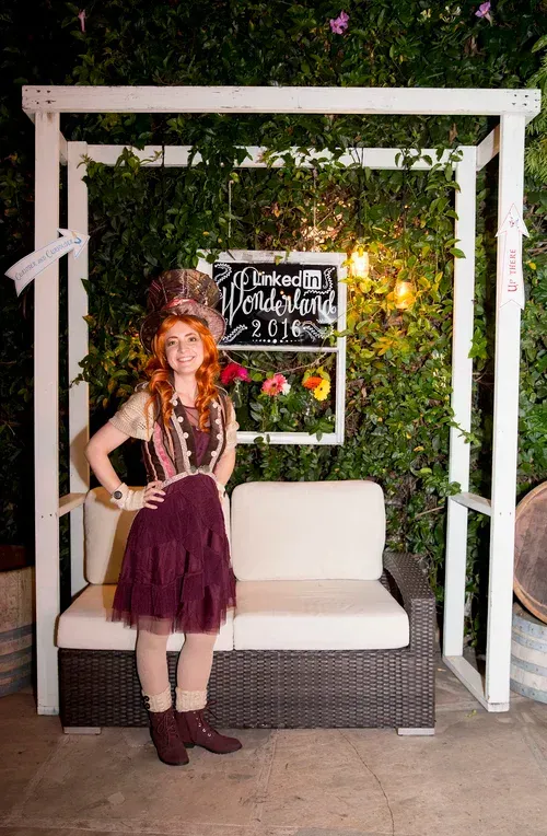 Woman posing beside a white outdoor sofa under a pergola with greenery and a chalkboard sign lit by fairy lights