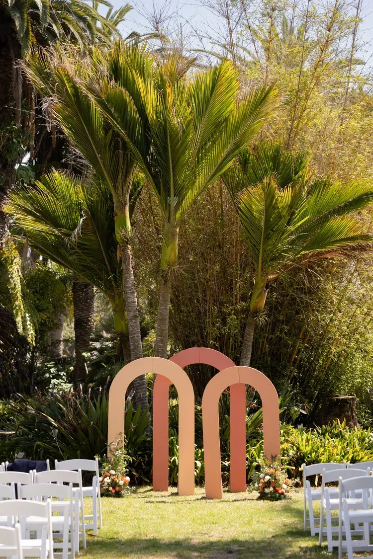 White chairs on grass facing a pink arch sculpture amid lush tropical greenery outdoors