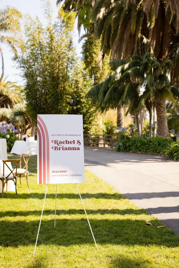 Event sign on a lawn beside a sunlit garden path with palm trees and chairs.