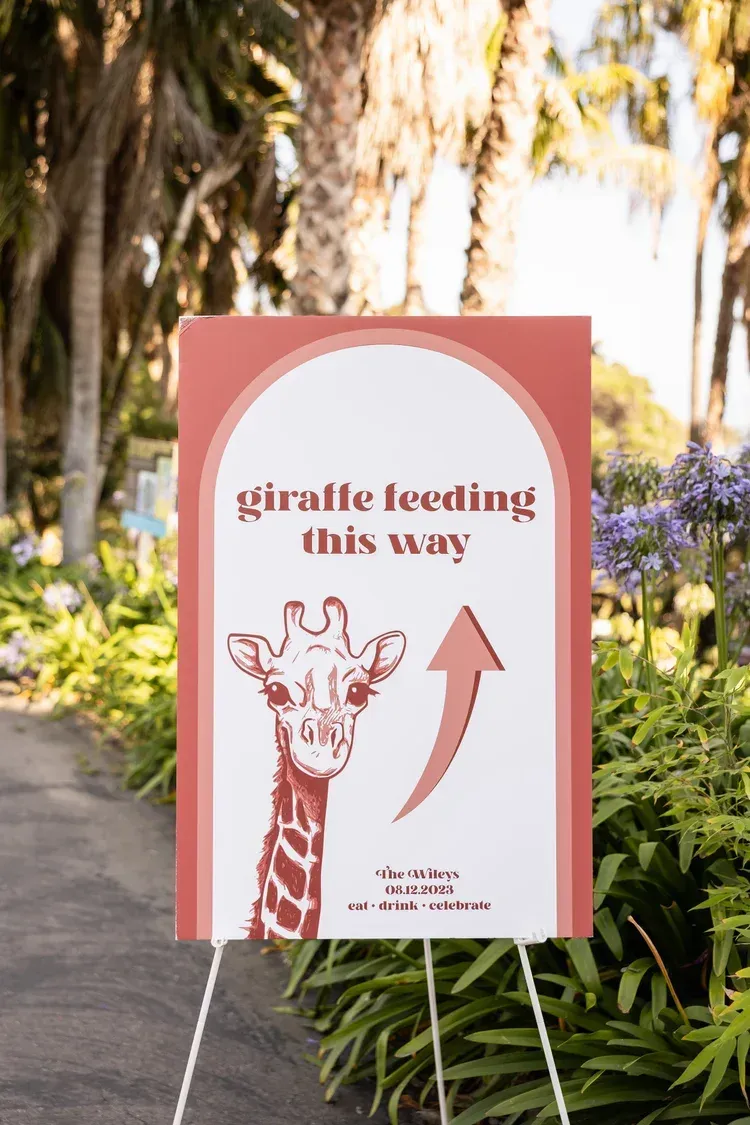 Sign pointing to giraffe feeding, with giraffe illustration and arrow, in a garden setting