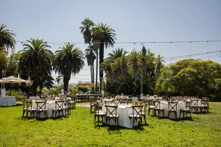 Outdoor garden event setup with round tables and white chairs on a grassy lawn under palm trees