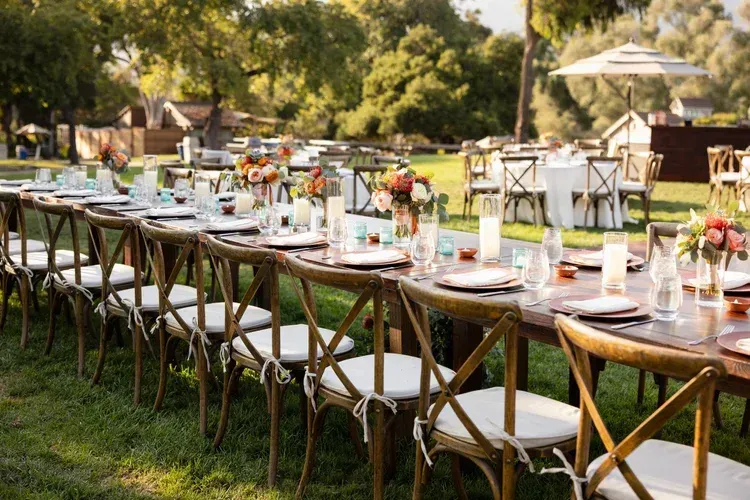 Long outdoor banquet table set with white tablecloths, glassware, and floral centerpieces in a garden