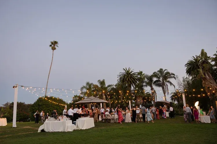 Outdoor evening garden reception with string lights, white tables, and guests mingling on a lawn.