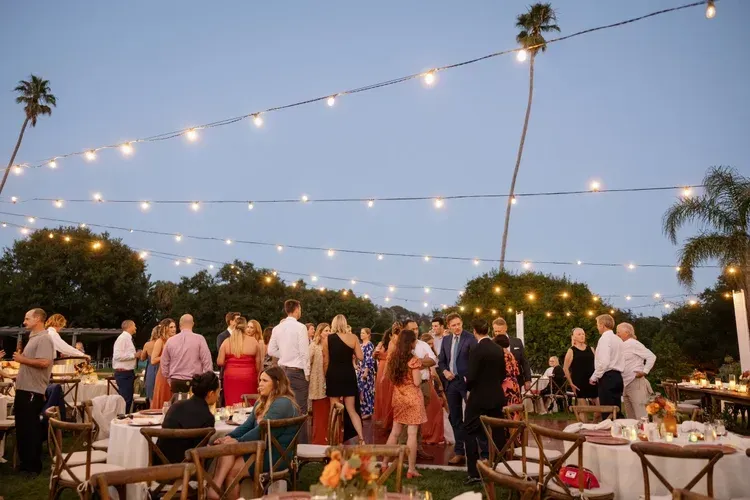 Outdoor evening party with string lights, guests mingling around tables at sunset