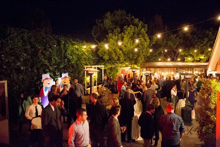 Crowded outdoor evening party with string lights, trees, and guests mingling near a lit building