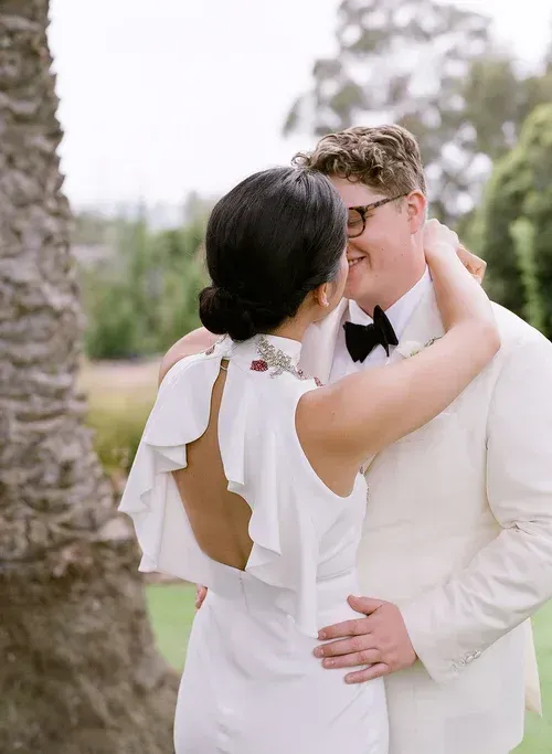 Bride and groom embracing outdoors, she in a white dress with open back, he in a white suit and black bow tie.