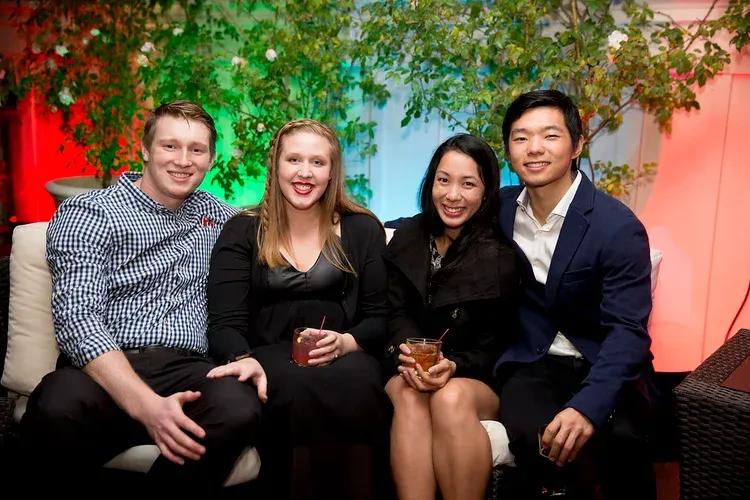 Four people sit on a couch, smiling, with greenery and warm red lighting in the background.
